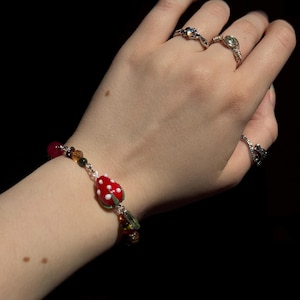 Könnte beinhalten: Eine Hand mit drei Ringen und einem Armband. Die Ringe sind silberfarben, jeder mit einem anderen Edelstein. Das Armband hat einen rot-weißen Pilzanhänger und rote Perlen. Der Hintergrund ist schwarz.