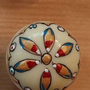 May include: A cream-coloured, spherical decorative object with a floral design. The design features gold, red, and blue petal-shaped elements, outlined in black. Small red, blue, and green accents are scattered around the sphere.
