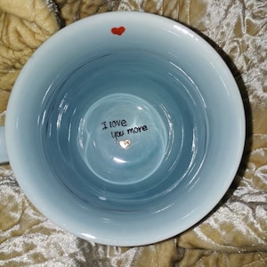 May include: A light blue ceramic mug with a white rim. The interior features the handwritten text "I love you more" with a small heart. A red heart is painted near the rim.
