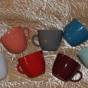May include: A collection of seven ceramic mugs in various colors, including coral, gray, teal, light blue, red, burgundy, and light gray. Each mug has a rounded shape and a handle.