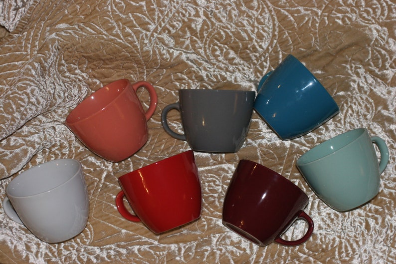 May include: A collection of seven ceramic mugs in various colors, including coral, gray, teal, light blue, red, burgundy, and light gray. Each mug has a rounded shape and a handle.