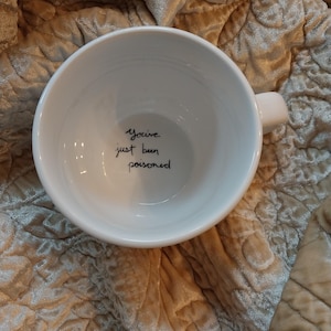 May include: A white ceramic mug with a handle, featuring the handwritten phrase "You've just been poisoned" inside. The mug is set against a textured, quilted fabric background.