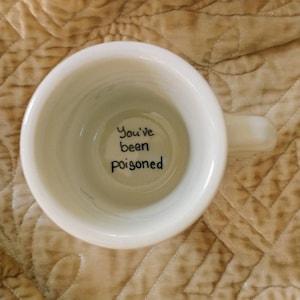 May include: White ceramic mug with a handle, viewed from above, revealing the words "You've been poisoned" in black script inside the mug. The mug is set against a textured, tan background.