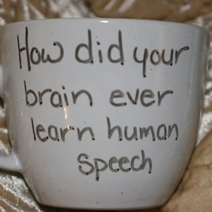 May include: White ceramic mug with a curved handle. The mug features the handwritten text: "How did your brain ever learn human speech?" The mug is set against a textured, golden-colored background.