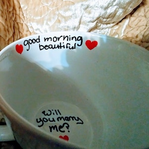 Puede incluir: Taza de cerámica blanca con las frases "good morning beautiful" y "Will you marry me?" escritas en letra negra. Acentos de corazón rojo se colocan cerca del texto. La taza tiene un asa curva.
