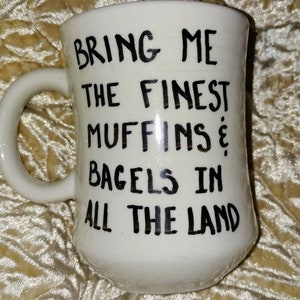 May include: White ceramic mug with a curved handle. The mug features black handwritten text that reads "BRING ME THE FINEST MUFFINS & BAGELS IN ALL THE LAND."