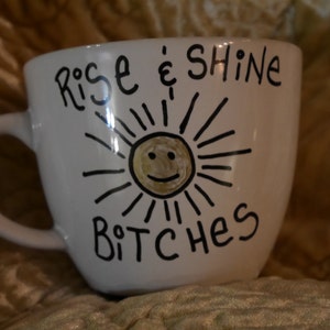 May include: White ceramic mug with a black handle, featuring the phrase "Rise & Shine Bitches" in black script. A smiling sun with radiating lines is centered below the text. The mug is on a textured, golden-brown surface.