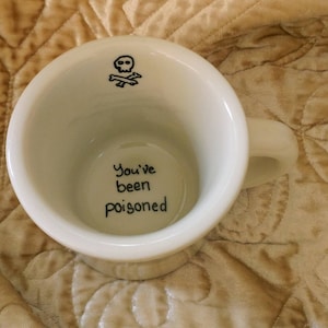 May include: White ceramic mug with a handle, featuring a black skull and crossbones graphic inside the rim. The words "You've been poisoned" are written in black script at the bottom of the mug.