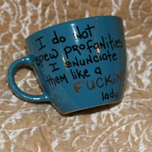 May include: A teal ceramic mug with a handle, featuring the handwritten phrase "I do not spew profanities I enunciate them like a FUCKING lady" in black and copper lettering.