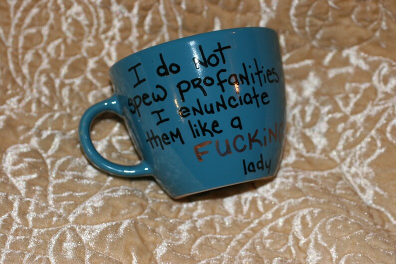 May include: A teal ceramic mug with a handle, featuring the handwritten phrase "I do not spew profanities I enunciate them like a FUCKING lady" in black and copper lettering.