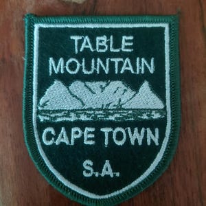 May include: A shield-shaped embroidered patch in dark green with a white border. It features white stitched text reading TABLE MOUNTAIN, CAPE TOWN, and S.A., along with a white embroidered graphic of the Table Mountain range in South Africa.