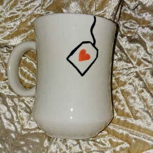 May include: White ceramic mug with a curved handle. The mug features a black outline of a tea bag with a red heart inside. The mug has a glossy finish and is designed for hot beverages.
