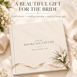 Könnte beinhalten: Ein quadratisches, weißes Hardcover-Buch mit dem aufgedruckten Text "OPEN ME BEFORE YOU SAY I DO" und "Madison • September 17, 2026". Das Cover zeigt eine Grafik eines Calla-Lilien-Straußes mit einer Schleife sowie dezente, wellenförmige Linien.