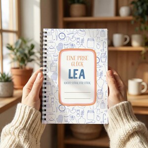 May include: A spiral-bound notebook with a white cover featuring a blue line-art pattern of kitchen utensils and jars. A central orange-bordered label contains the text: EINE PRISE GLÜCK LEA KOCHT STÜCK FÜR STÜCK, with two blank lines below.