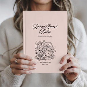 Könnte beinhalten: Ein hellrosa Baby-Journal mit dem Text "Berry Sweet Baby" und einer Schwarz-Weiß-Illustration von Erdbeeren in einem Korb. Das Journal enthält auch den Text "Growing Sweeter Every Day" und "Celebrating Baby Emily July 23, 2020."