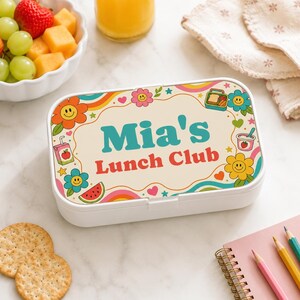 Könnte beinhalten: Eine rechteckige weiße Brotdose mit einem cremefarbenen Deckel, der den Schriftzug Mia's Lunch Club in Türkis und Rot trägt. Das Design zeigt bunte Retro-Blumen mit lächelnden Gesichtern, Herzen, Sternen, eine Saftpackung und eine Wassermelone.