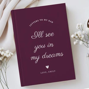 Könnte beinhalten: Ein tiefburgunderfarbenes Notizbuch mit festem Einband, das oben den weißen Aufdruck "LETTERS TO MY DAD" trägt, gefolgt von dem kursiven Schriftzug "I'll see you in my dreams", einem kleinen weißen Herzsymbol und der Signatur "LOVE, EMILY" unten.