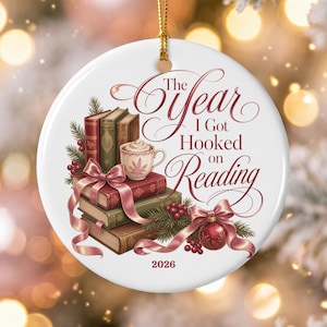 Könnte beinhalten: Ein rundes, weißes Keramikornament mit einem Stapel Bücher, einer Tasse und einer roten Weihnachtskugel, die mit einem rosa Band verziert ist. Der Text lautet The Year I Got Hooked on Reading und 2026. Das Motiv zeigt Tannenzweige und rote Beeren.