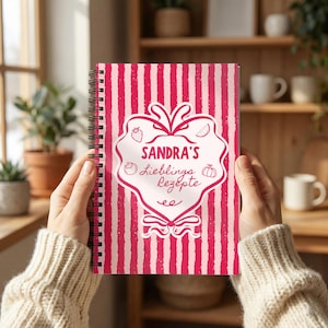 May include: A spiral-bound notebook with a pink and white vertical striped cover. The centre features a white heart-shaped graphic with the text "SANDRA'S Lieblins Rezepte" and small line drawings of a strawberry, a slice of melon, a tomato, and a pumpkin.
