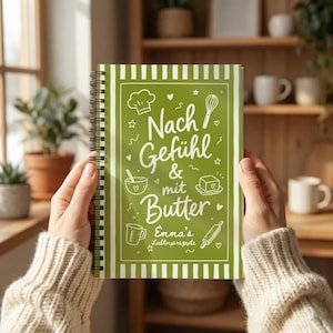 Könnte beinhalten: Ein spiralgebundenes Notizbuch mit grünem Einband, weißen Streifenrändern und weißen Strichzeichnungen von Küchenutensilien. Der Text lautet "Nach Gefühl & mit Butter" sowie "Emma's Lieblingsrezepte" in einer weißen Schreibschrift.