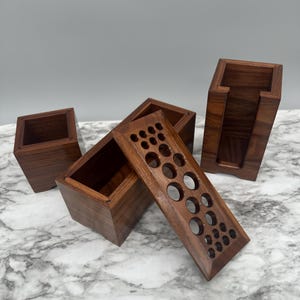 May include: Set of four wooden boxes with different designs. One box has a lid with a pattern of circles, another box has a lid with a pattern of squares, and the other two boxes are plain. The boxes are made of dark wood and have a natural finish.