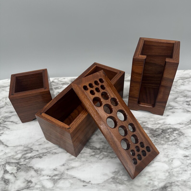 May include: Set of four wooden boxes with different designs. One box has a lid with a pattern of circles, another box has a lid with a pattern of squares, and the other two boxes are plain. The boxes are made of dark wood and have a natural finish.