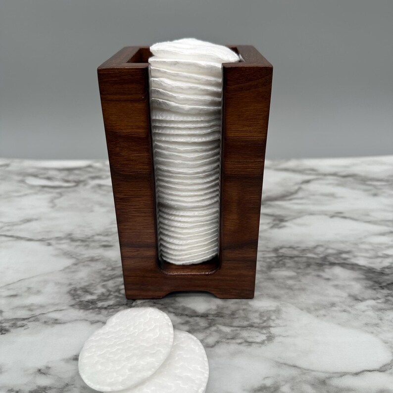 May include: A dark brown wooden cotton round storage container with a stack of white cotton rounds inside.