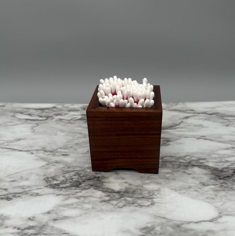 May include: A dark brown wooden box with a square shape and rounded edges, filled with white cotton swabs.