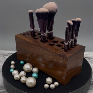 May include: A wooden makeup brush holder with a dark brown finish. The holder has multiple slots to store brushes of different sizes. The holder is filled with a variety of makeup brushes, including a large powder brush, a blush brush, and several smaller brushes.
