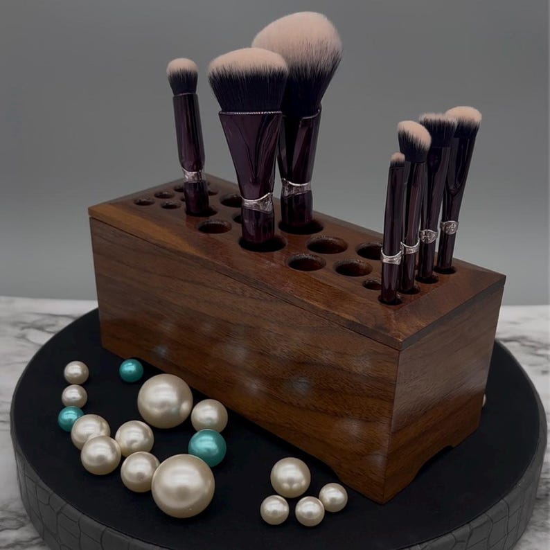 May include: A wooden makeup brush holder with a dark brown finish. The holder has multiple slots to store brushes of different sizes. The holder is filled with a variety of makeup brushes, including a large powder brush, a blush brush, and several smaller brushes.
