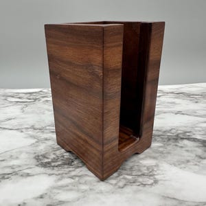 May include: A wooden napkin holder made of dark walnut wood. The holder has a rectangular shape with a slot in the front for holding napkins.