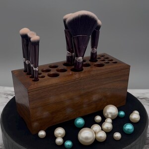 May include: A dark brown wooden makeup brush holder with multiple holes to hold brushes. The holder is filled with various makeup brushes, including a large fan brush with black and pink bristles. The holder is sitting on a black surface with white and teal pearls scattered around it.