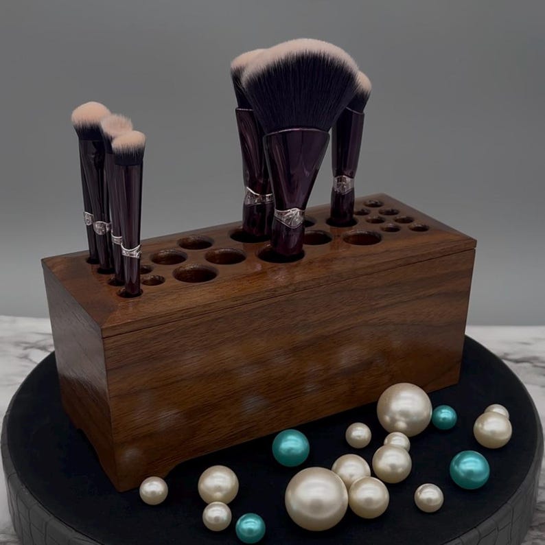 May include: A dark brown wooden makeup brush holder with multiple holes to hold brushes. The holder is filled with various makeup brushes, including a large fan brush with black and pink bristles. The holder is sitting on a black surface with white and teal pearls scattered around it.