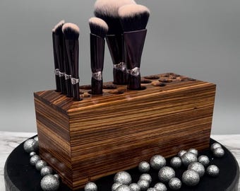 May include: A wooden makeup brush holder with a striped wood grain pattern. The holder has multiple slots to hold brushes upright. There are seven makeup brushes in the holder, including a large fluffy brush.