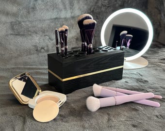 May include: A black wooden makeup brush holder with gold trim, filled with various makeup brushes. A compact mirror, powder compact with a sponge, and two lavender makeup brushes are also present. A lighted vanity mirror is in the background.