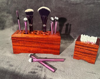 May include: A wooden makeup brush holder with various brushes and a small wooden container with cotton swabs. The brush holder is a reddish-brown color and has several holes to hold the brushes. The brushes have purple handles and white bristles.