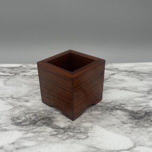 May include: A small, square wooden planter box with a dark brown stain. The planter has a simple design and is perfect for small plants or succulents.