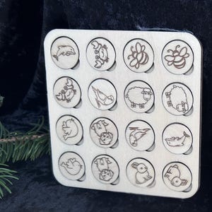 Könnte beinhalten: Ein Holzpuzzlespiel mit 16 kreisförmigen Aussparungen, die jeweils eine lasergravierte Tierillustration enthalten. Zu den Tieren gehören ein Delfin, eine Krabbe, eine Biene, ein Schaf und ein Vogel. Das Brett ist aus hellem Holz gefertigt.