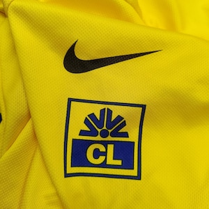 Puede incluir: Camiseta deportiva amarilla brillante con un logotipo de Nike negro y un logotipo azul y amarillo. El logotipo es un cuadrado con las letras "CL" en su interior. La camiseta est&aacute; hecha de una tela texturizada y transpirable.
