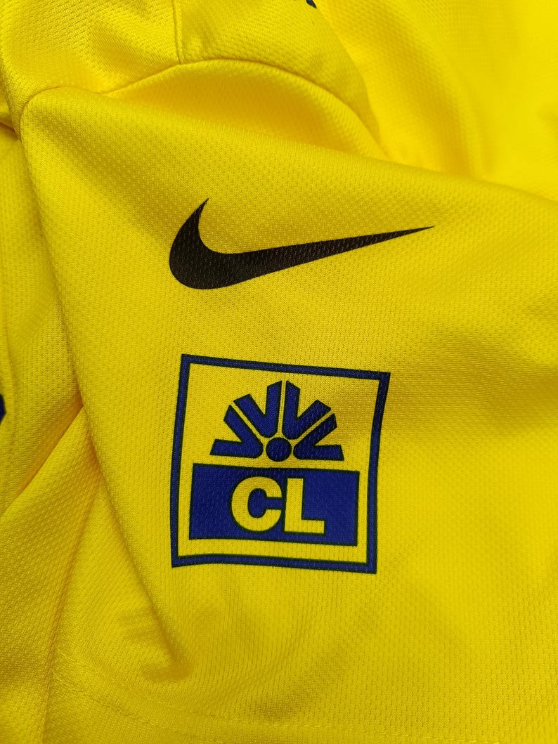 Puede incluir: Camiseta deportiva amarilla brillante con un logotipo de Nike negro y un logotipo azul y amarillo. El logotipo es un cuadrado con las letras "CL" en su interior. La camiseta est&aacute; hecha de una tela texturizada y transpirable.