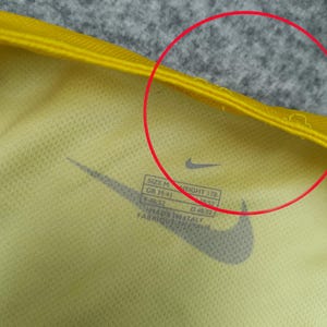 Puede incluir: Primer plano de una camiseta deportiva amarilla con el logotipo negro de Nike. La camiseta tiene una etiqueta con informaci&oacute;n de talla y medidas. La tela es de color amarillo claro.
