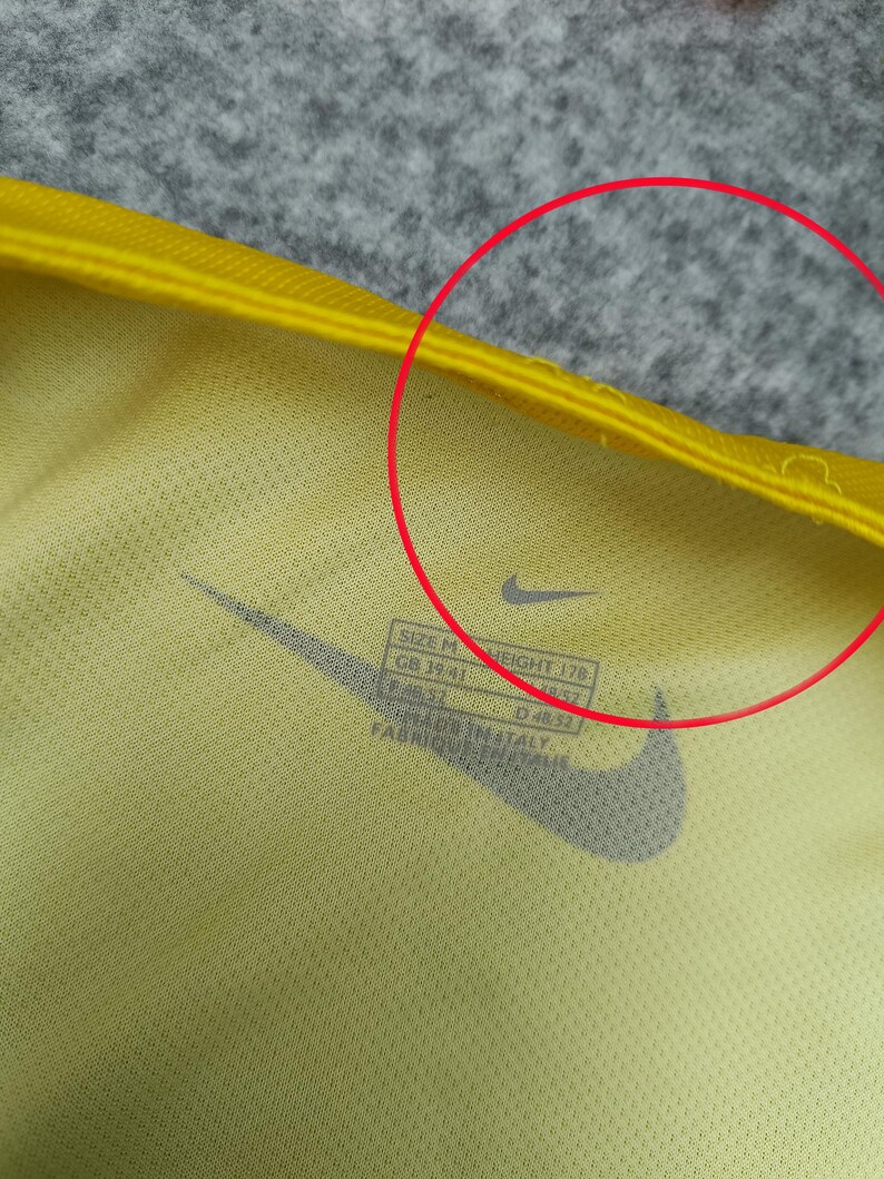 Puede incluir: Primer plano de una camiseta deportiva amarilla con el logotipo negro de Nike. La camiseta tiene una etiqueta con informaci&oacute;n de talla y medidas. La tela es de color amarillo claro.