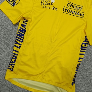 Puede incluir: Maillot de ciclismo amarillo con texto y logotipos azules. Presenta la marca "Le Tour de France 2005" y "Credit Lyonnais". Incluye un logotipo de Nike y cierre de cremallera. El maillot tiene mangas cortas.