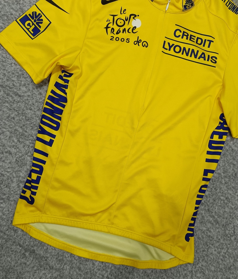 Puede incluir: Maillot de ciclismo amarillo con texto y logotipos azules. Presenta la marca "Le Tour de France 2005" y "Credit Lyonnais". Incluye un logotipo de Nike y cierre de cremallera. El maillot tiene mangas cortas.