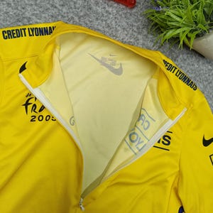 Puede incluir: Camiseta deportiva amarilla con cremallera parcial, con el texto "CREDIT LYONNAIS" en el hombro y "de Fra 2005" en el pecho. Un logotipo de Nike es visible en el interior del cuello. La camiseta est&aacute; hecha de tejido ligero y transpirable.