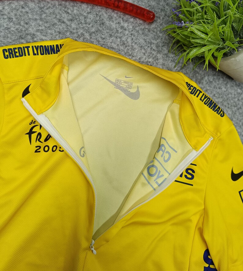 Puede incluir: Camiseta deportiva amarilla con cremallera parcial, con el texto "CREDIT LYONNAIS" en el hombro y "de Fra 2005" en el pecho. Un logotipo de Nike es visible en el interior del cuello. La camiseta est&aacute; hecha de tejido ligero y transpirable.