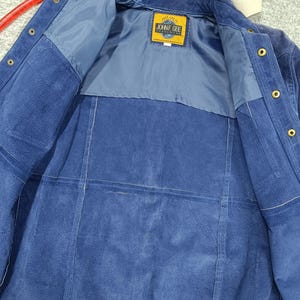 Peut inclure: Veste en daim bleu avec une fermeture boutonn&eacute;e et un col. La doublure int&eacute;rieure est d'une nuance de bleu plus claire. L'&eacute;tiquette indique "JOHN F. GEE". La veste est suspendue &agrave; un cintre rouge.