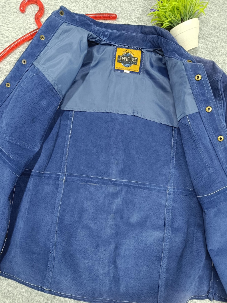 Peut inclure: Veste en daim bleu avec une fermeture boutonn&eacute;e et un col. La doublure int&eacute;rieure est d'une nuance de bleu plus claire. L'&eacute;tiquette indique "JOHN F. GEE". La veste est suspendue &agrave; un cintre rouge.