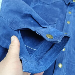 Peut inclure: Gros plan sur une chemise en daim bleu. La chemise a une fermeture boutonn&eacute;e sur le devant, deux poches de poitrine et des poignets boutonn&eacute;s. Les boutons sont dor&eacute;s. Le tissu a un aspect doux et textur&eacute;.