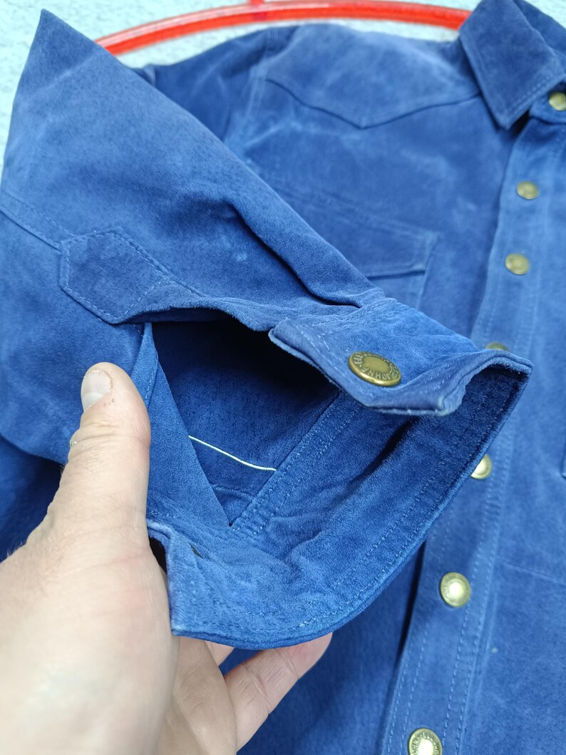 Peut inclure: Gros plan sur une chemise en daim bleu. La chemise a une fermeture boutonn&eacute;e sur le devant, deux poches de poitrine et des poignets boutonn&eacute;s. Les boutons sont dor&eacute;s. Le tissu a un aspect doux et textur&eacute;.